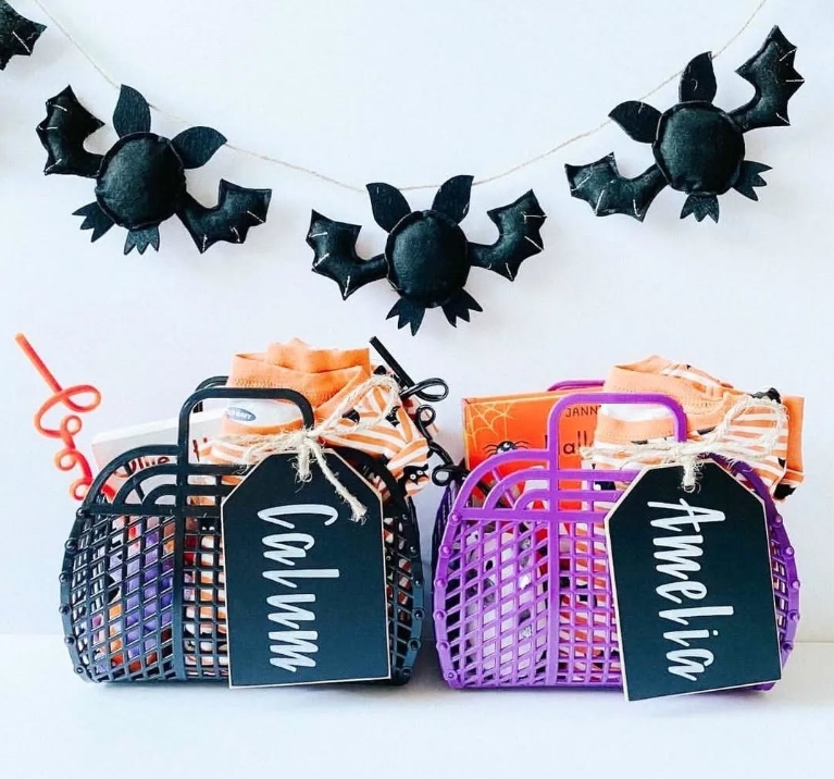 Two personalized Halloween baskets in black and purple, labeled 'Calum' and 'Amelia', filled with festive goodies and adorned with a bat garland in the background.