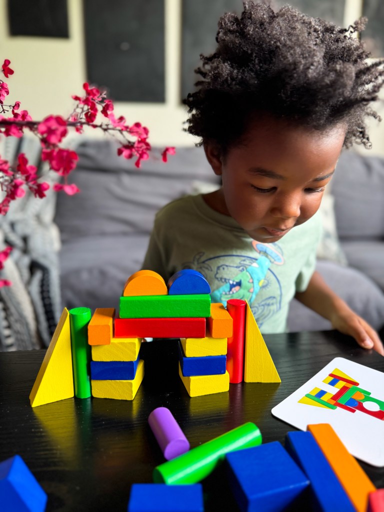 A young child focused on playing with colorful building blocks on a table, surrounded by loose blocks and a card showing building designs.
