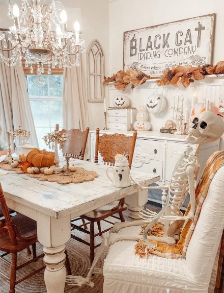 A cozy dining room decorated for fall, featuring a skeleton seated at a white table surrounded by pumpkins and autumn decor. The space is illuminated by a chandelier and decorated with a sign that reads 'Black Cat Trading Company'.