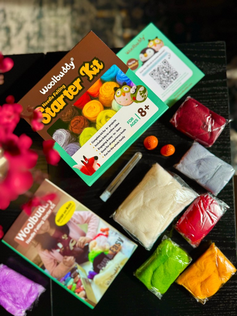 A Woolbuddy Needle Felting Starter Kit displayed on a table, showcasing colorful wool roving, felting needles, a foam pad, and an instruction booklet, surrounded by vibrant packaging.