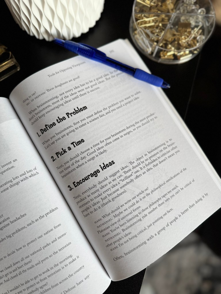 An open book displaying a page with instructions on brainstorming, including steps like 'Define the Problem,' 'Pick a Time,' and 'Encourage Ideas,' alongside a blue pen and a white decorative vase in the background.
