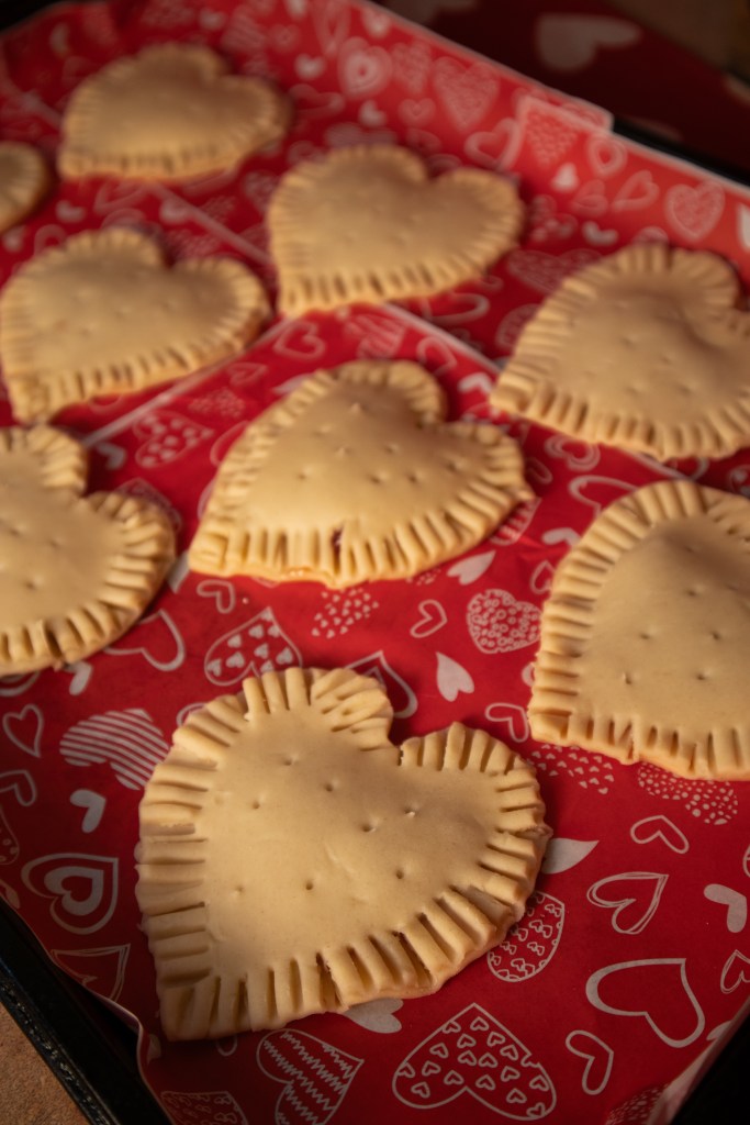 A tray of uncooked heart-shaped pop tarts on red parchment paper decorated with hearts.