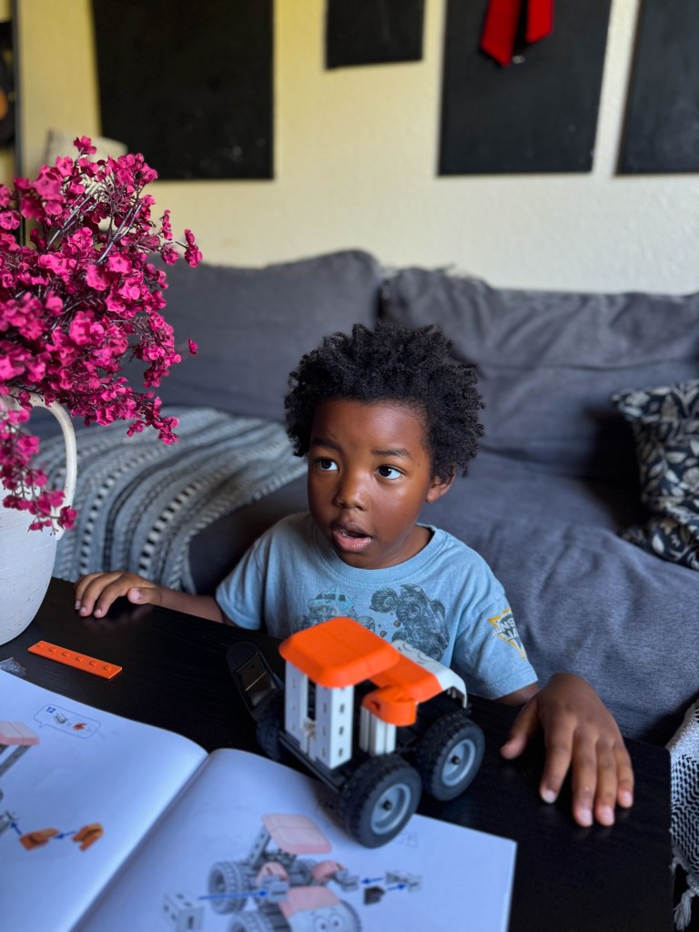 A young boy with curly hair is sitting at a table, looking surprised while playing with a toy construction vehicle. A building instruction manual and a decorative flower arrangement are visible on the table.
