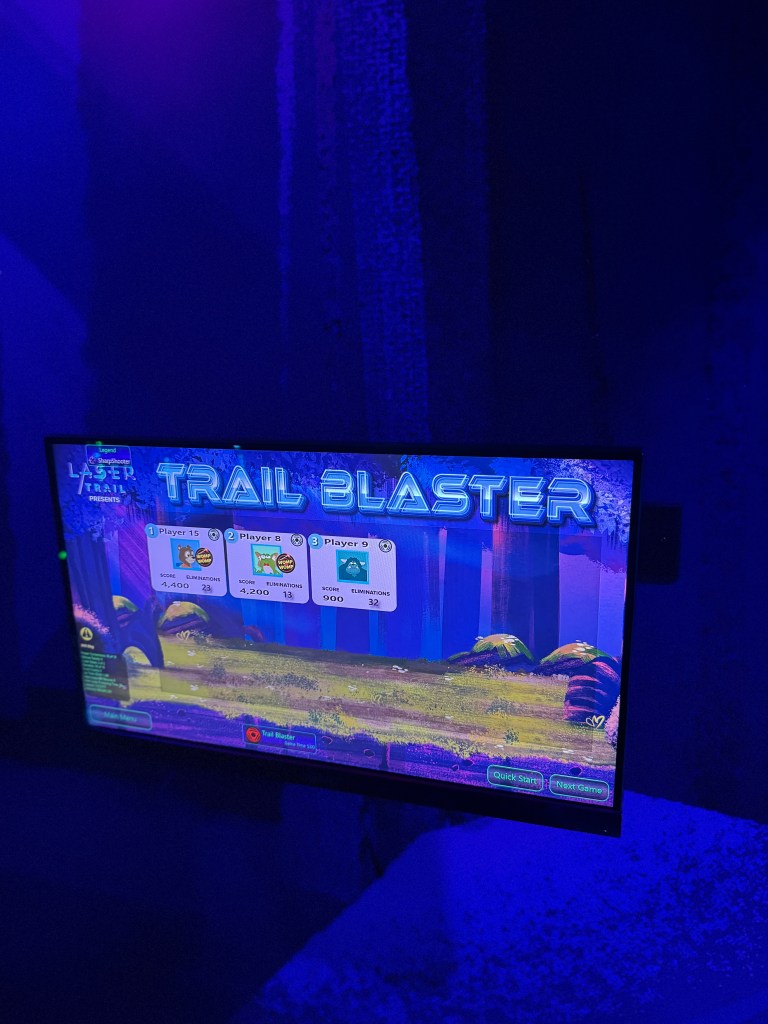 A digital screen displaying the 'Trail Blaster' game interface in a darkened room with blue lighting, showing player scores and statistics.