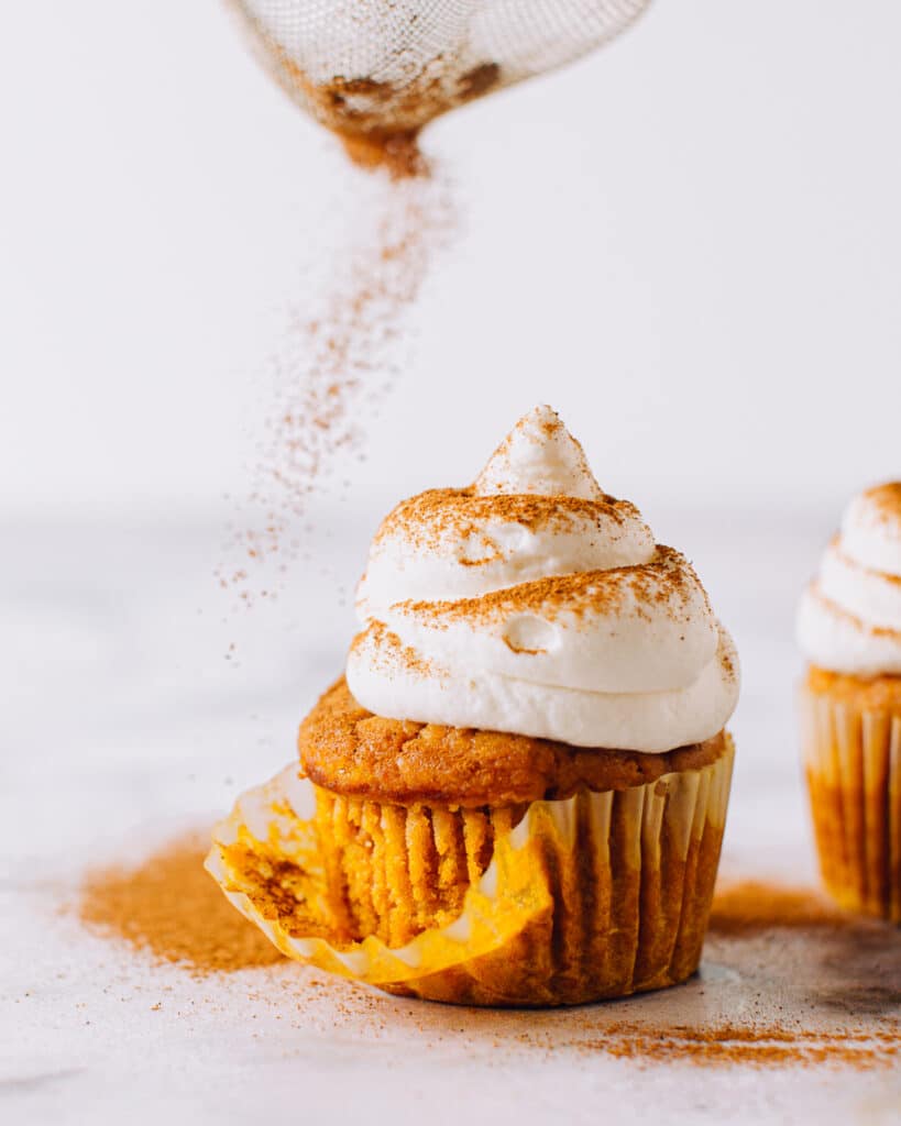 A close-up of a pumpkin cupcake with a swirl of whipped cream frosting topped with cinnamon dust, sitting next to its removed cupcake liner.
