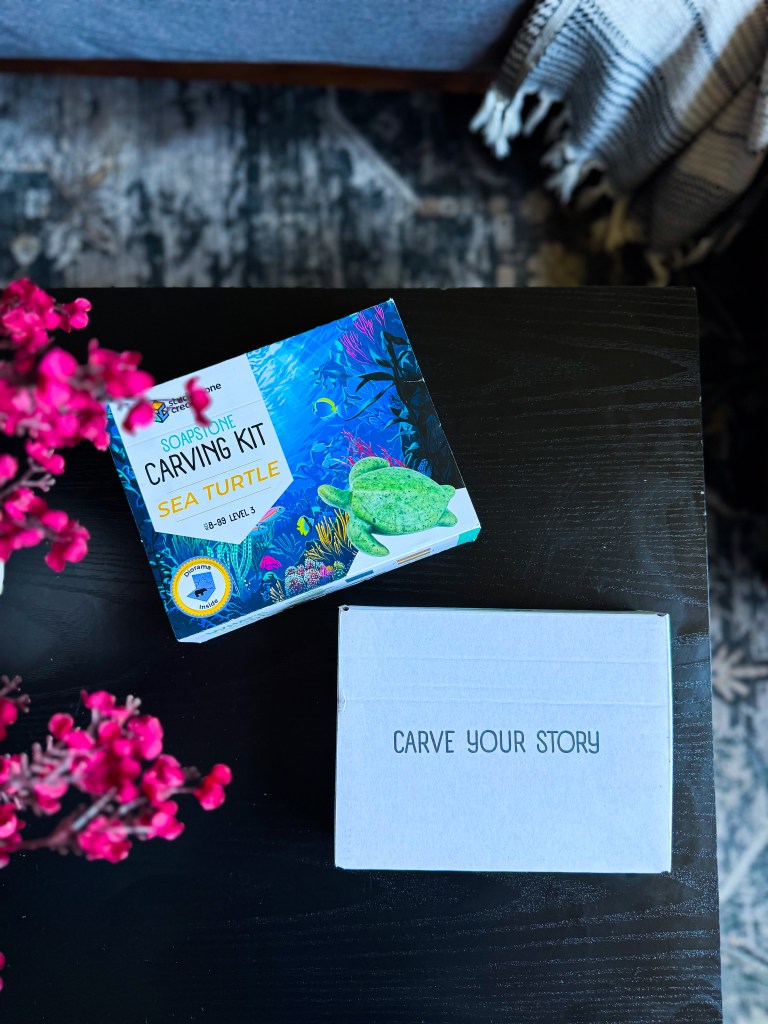 A soapstone carving kit featuring a sea turtle design, placed on a dark wooden table alongside a plain box that says 'CARVE YOUR STORY.' A touch of pink flowers is seen in the foreground.