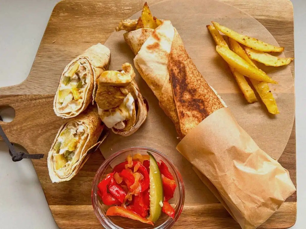 A wooden cutting board featuring a delicious meal with two rolled tortillas filled with chicken, cheese, and vegetables, alongside a wrapped larger tortilla and a side of golden fries. A small bowl of pickled peppers adds a colorful touch.