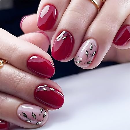 Close-up of manicured hands featuring a mix of deep red and nude nails with metallic leaf accents, showcasing a stylish fall nail design.