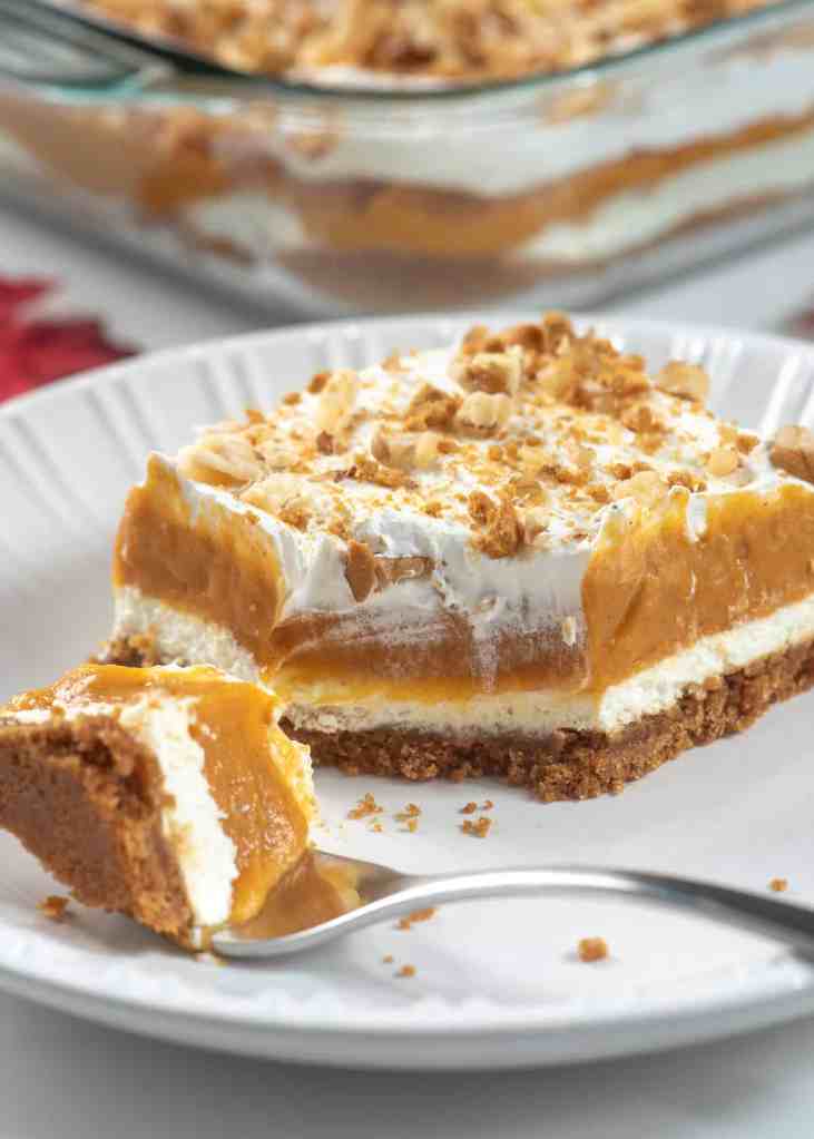 A slice of layered pumpkin dessert with a creamy filling and crushed nuts on top, served on a white plate with a fork.