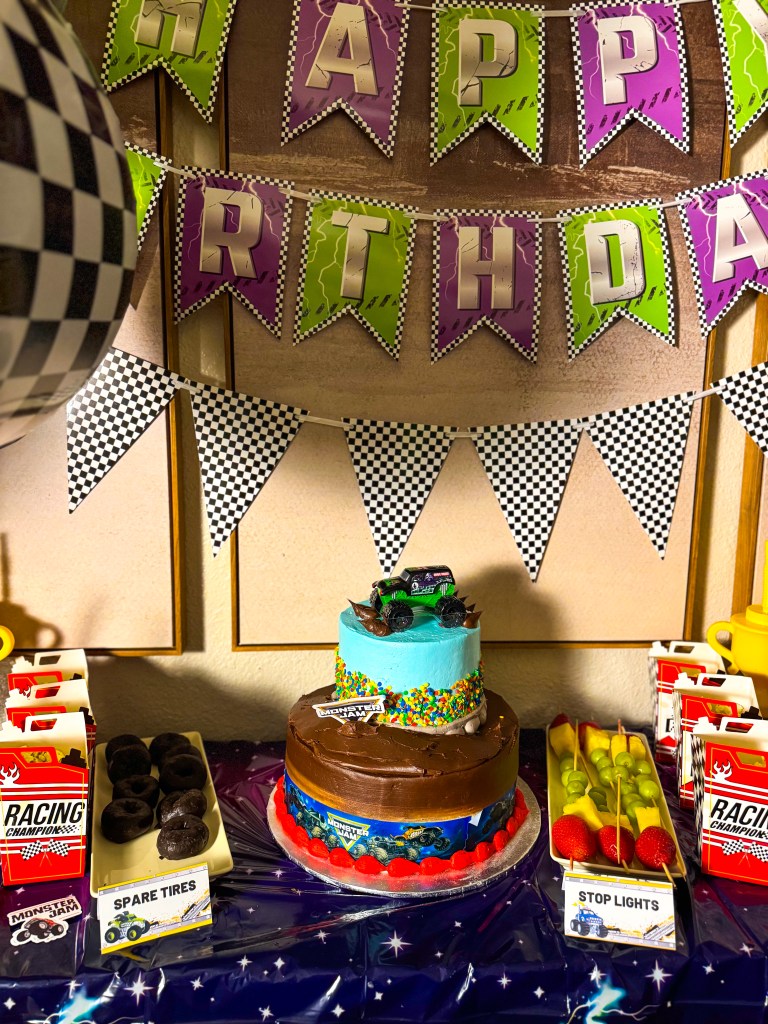 Monster Jam birthday cake with monster truck topper on a racing themed dessert table with party decorations and snacks.