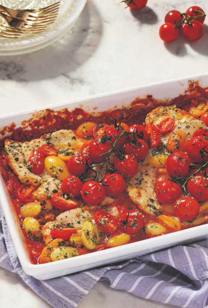 Baked chicken breast topped with cherry tomatoes in a white dish, garnished with herbs, resting on a striped cloth.