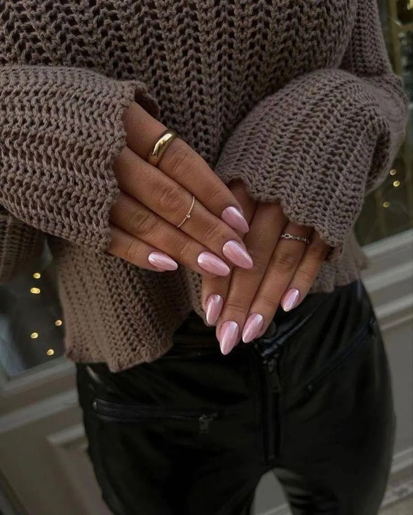 A close-up of hands displaying manicured pink nails, adorned with multiple rings, while wearing a cozy, knitted brown sweater.