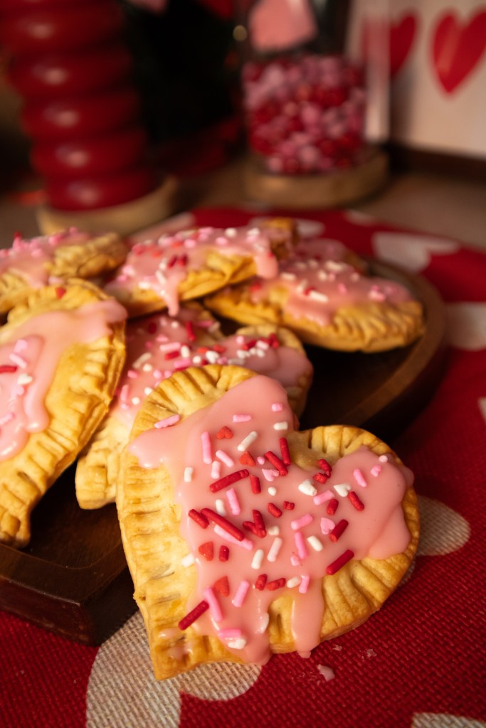 A close-up view of heart-shaped pop tarts topped with pink frosting and colorful sprinkles, presented on a wooden platter against a patterned background.