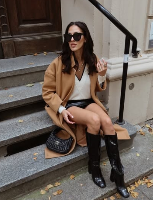 A woman sitting on steps wearing a camel coat, black knee-high boots, and a stylish outfit, with sunglasses and a patterned bag, surrounded by autumn leaves.