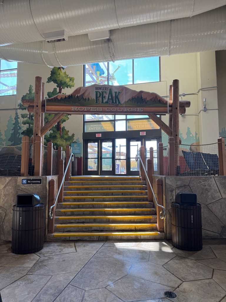 Entrance to Howlers Peak Ropes Course at Great Wolf Lodge with stairs leading up, surrounded by trees and indoor decorations.