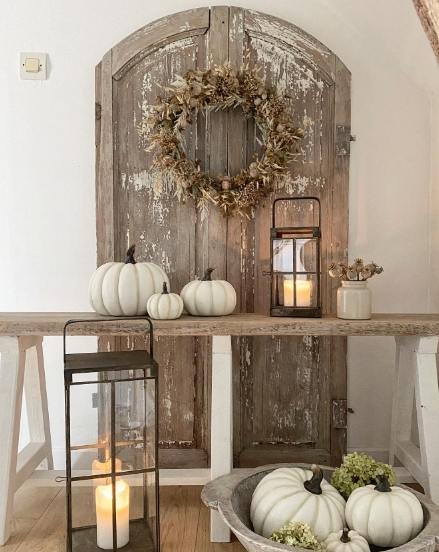A rustic wooden door with a wreath hangs above a wooden table displaying white pumpkins, a lantern, and a vase of dried flowers. The scene conveys a cozy autumn atmosphere.