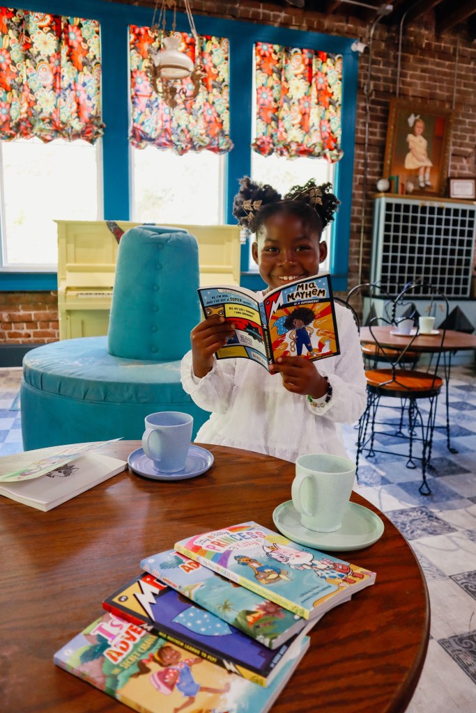 Child enjoying the Mia Mayhem superhero book with chapter books stacked on a nearby table.