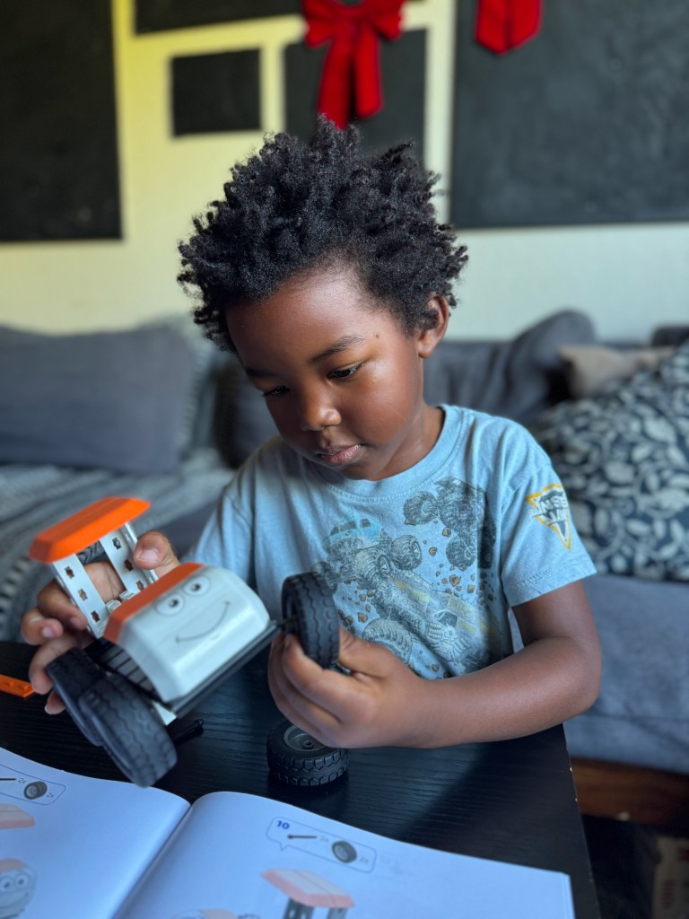 A young child with curly hair is assembling a toy vehicle, focused on attaching a wheel. The child is seated at a table with an instructional book open in front of them, in a cozy living room setting.