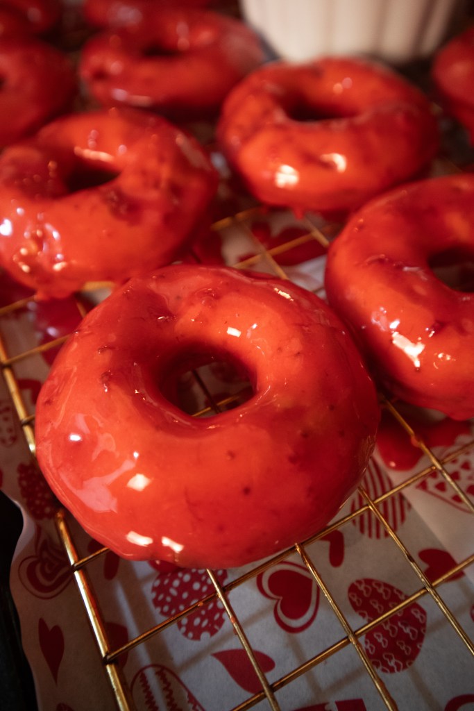 Close-up of freshly glazed red donuts on a cooling rack, with a decorative patterned background.