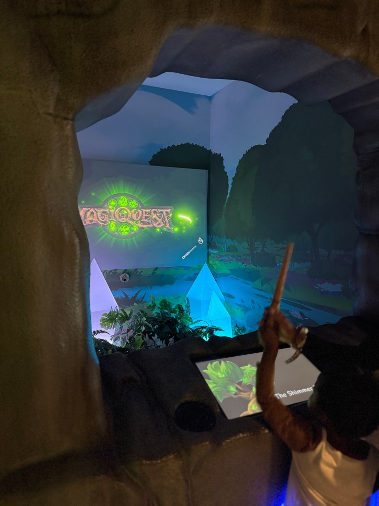 A child using a wand to activate a MagiQuest game in a themed adventure area, featuring vibrant graphics and props.