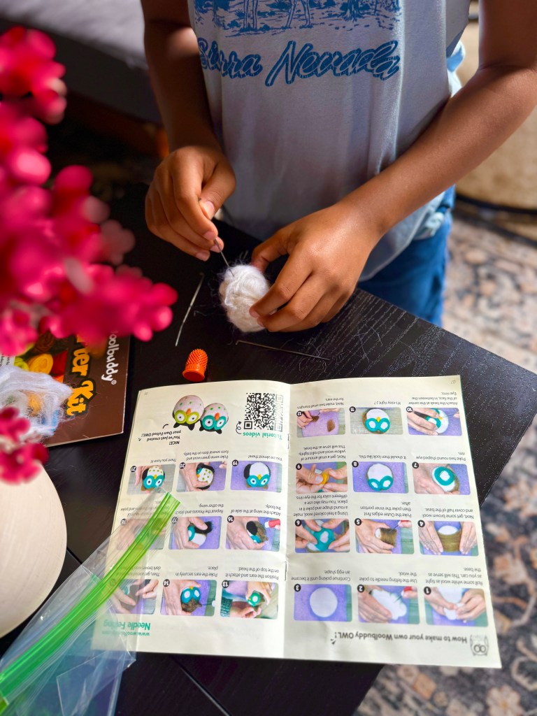 A child is working on a wool felting project using a felting needle and wool, with a colorful instruction booklet placed on the table showing steps to create felted owls.