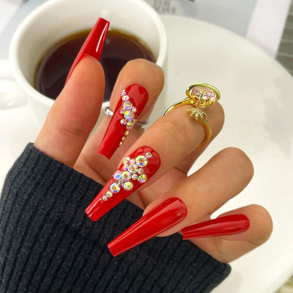 A close-up of manicured hands featuring long, glossy red nails with decorative rhinestones. A cup of coffee is in the background.