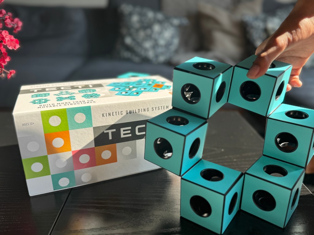 A hand is assembling a circular structure using turquoise cubes from the Tecti kinetic building system, with the packaging visible in the background.