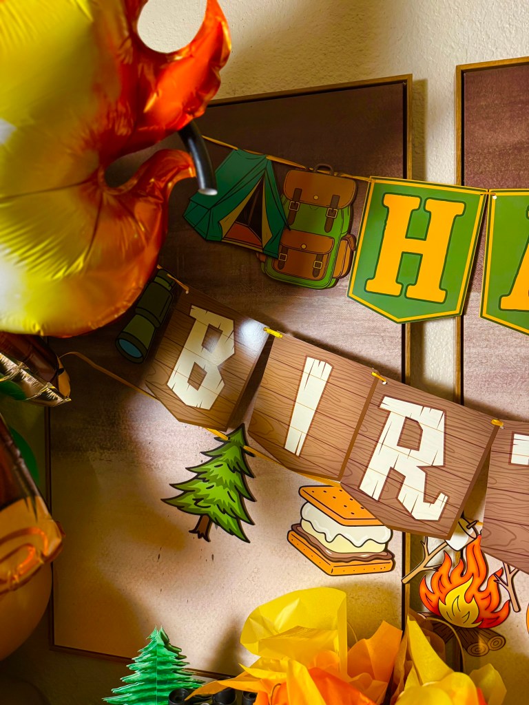 Close-up of camping birthday banner with tent, backpack, s’mores, bear, lantern, and campfire designs.