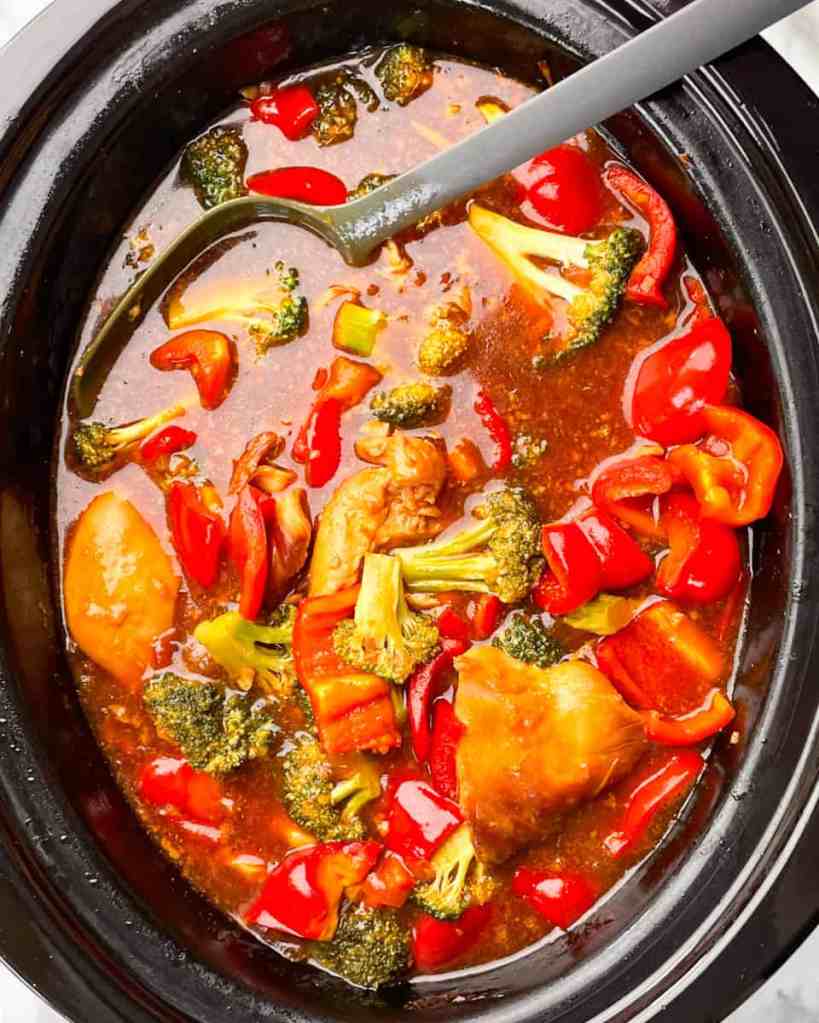A close-up view of a slow cooker filled with chicken and colorful vegetables, including red peppers and broccoli, simmering in a savory sauce.