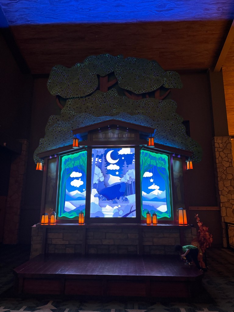 A beautifully lit lobby display at Great Wolf Lodge featuring a colorful forest scene with large trees and a moonlit background. The exhibit includes decorative lanterns and is designed to create an inviting atmosphere for families.