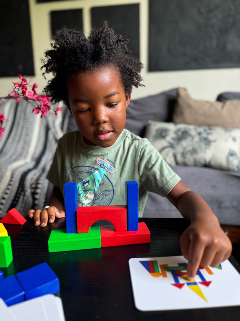 A child playing with colorful building blocks, constructing a structure on a table while looking at a card for guidance.