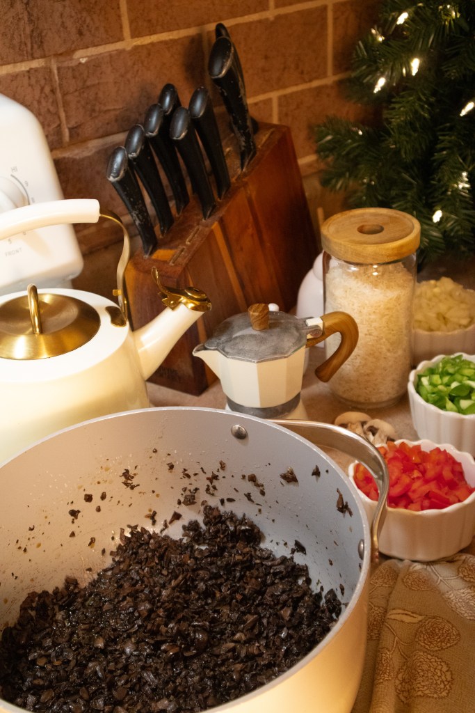 A kitchen scene featuring a pot with sautéed dark mushrooms, a white kettle, a knife block, and various chopped vegetables in bowls, including green onions and red bell peppers, with a festive touch of a decorated tree in the background.