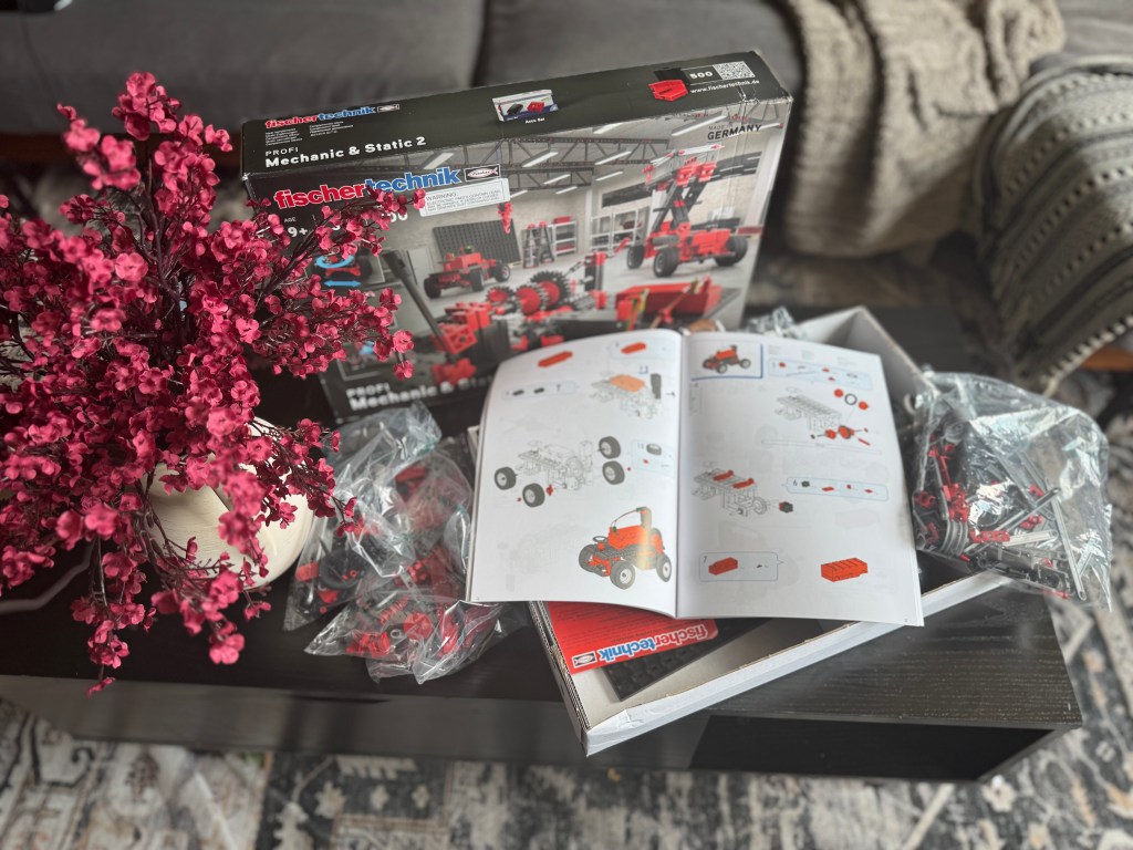 An opened Fischer Technik STEM kit box with a booklet and various building parts on a dark table, surrounded by a decorative plant.