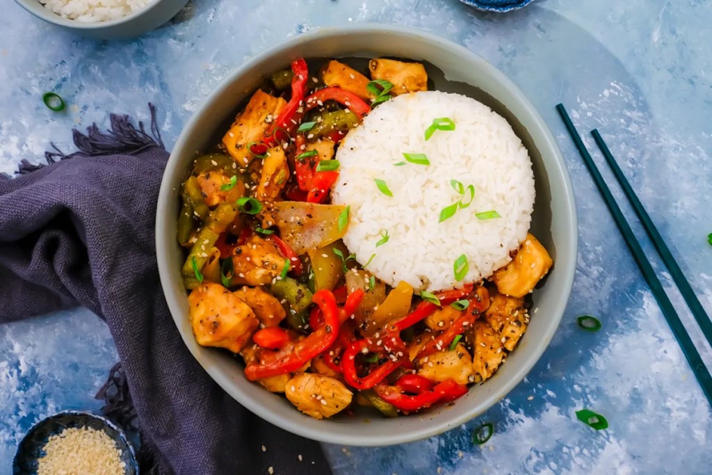 A bowl of colorful chicken stir fry with red and green bell peppers, onions, and sesame seeds, served with a portion of white rice and garnished with green onions.