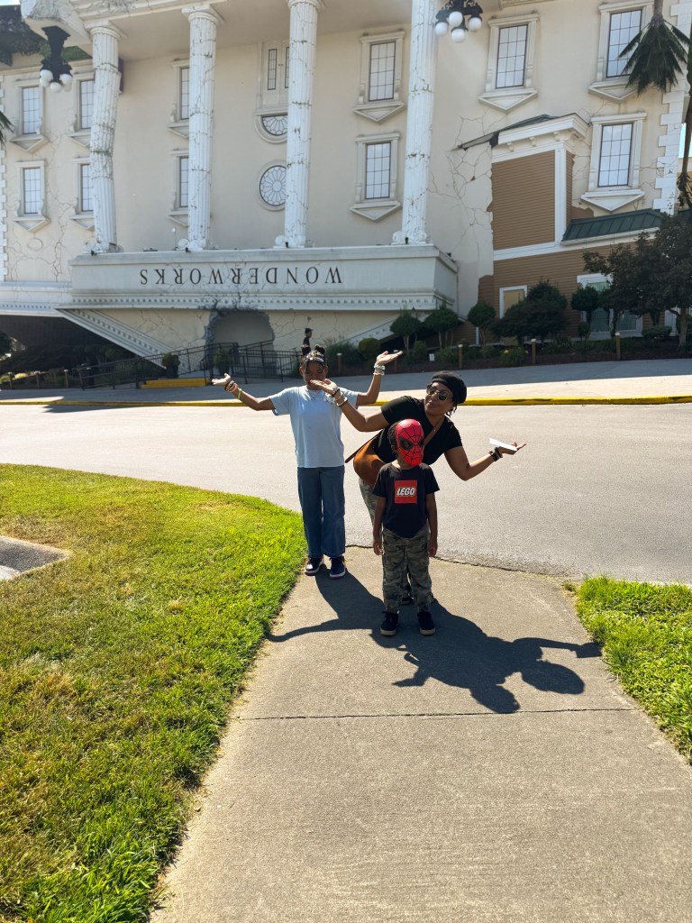 Three people posing playfully in front of an upside-down building called WonderWorks, with one child wearing a Spiderman mask.