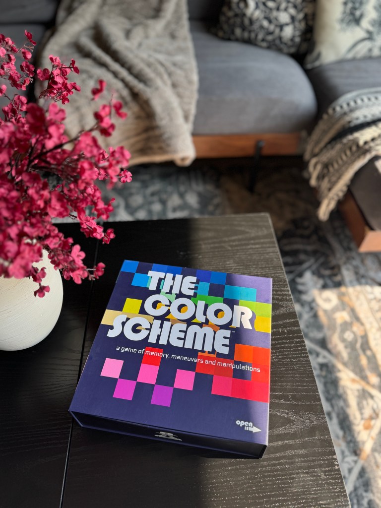 A closed box of 'The Color Scheme' board game on a dark wooden table, surrounded by a floral arrangement and a cozy sofa.