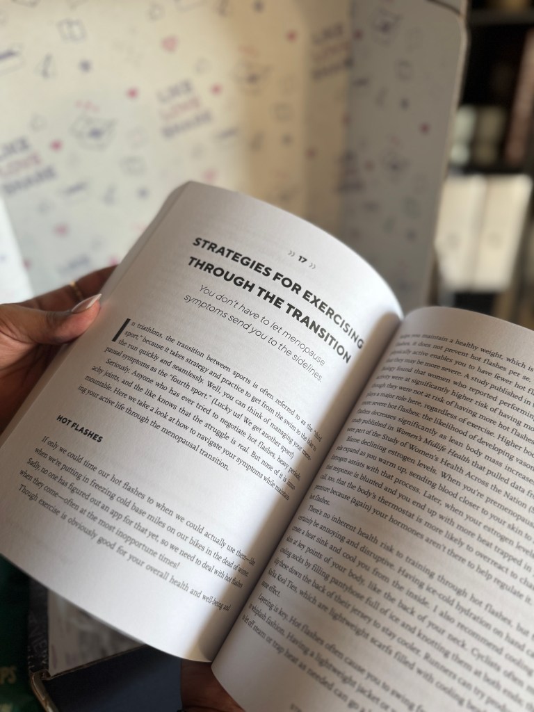 A close-up of a hand holding an open book with the heading 'Strategies for Exercising Through the Transition' on the page, discussing menopausal symptoms. The background features a softly decorated wall.
