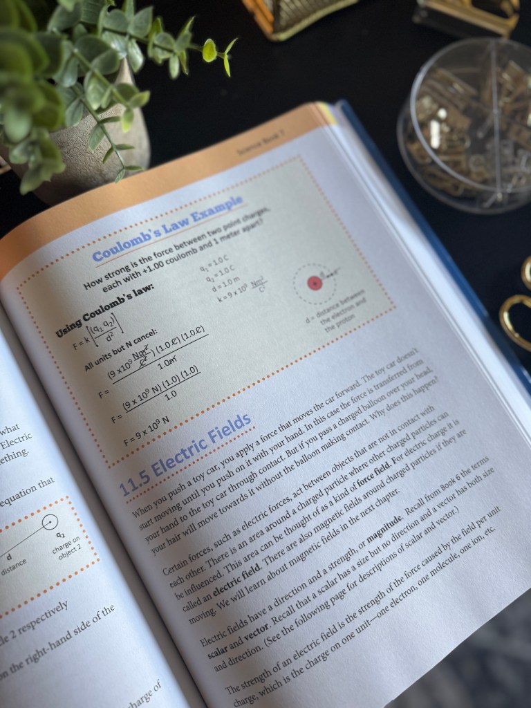 A close-up view of an open textbook page detailing Coulomb's Law and Electric Fields, showcasing illustrations and equations related to electrostatic forces, with a decorative plant and small objects visible in the background.