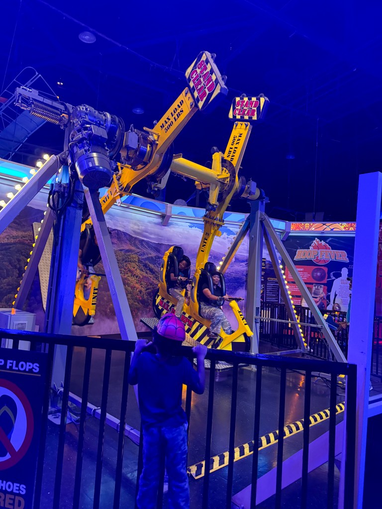 A thrilling amusement ride with a robotic arm holding seats for riders, set in a colorful indoor environment. A child watches from behind a safety barrier.