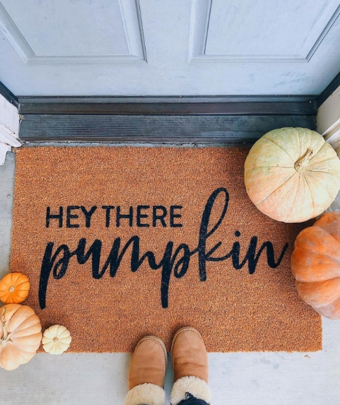 A cozy porch scene featuring a doormat with the playful greeting 'HEY THERE PUMPKIN' surrounded by a variety of pumpkins in warm fall colors.