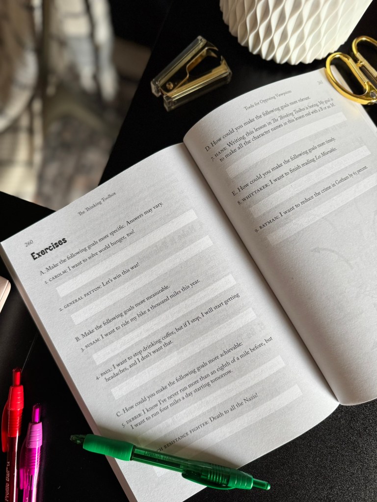 Open book displaying exercise pages from 'The Thinking Toolbox' with prompts for students, surrounded by pens and a stapler on a dark table.