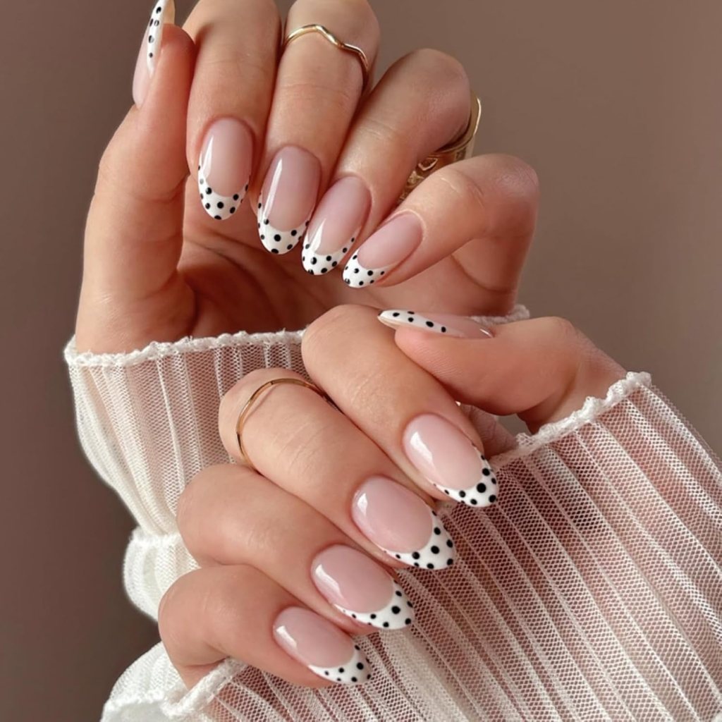 Close-up of a hand displaying manicured nails with a clear pink base and black polka dot tips, adorned with thin gold rings and a sheer white top.