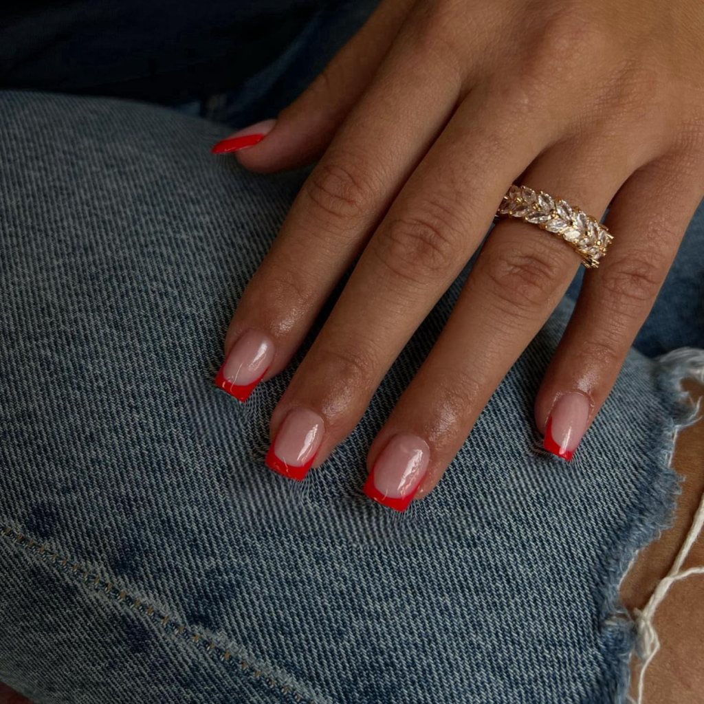 Close-up of a hand resting on denim fabric, showcasing a manicure with nude nails featuring red tips and a gold ring with clear stones.