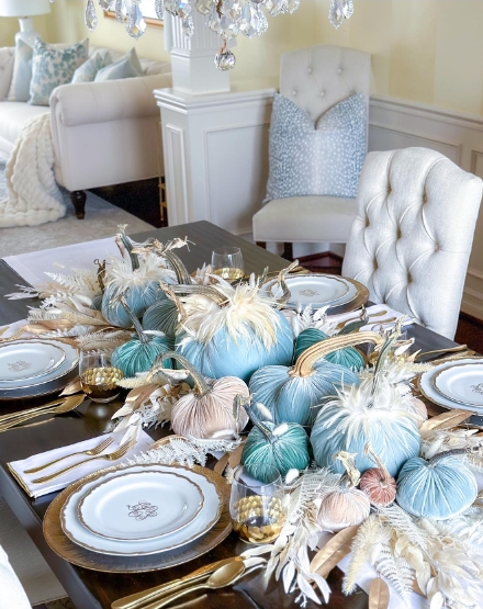 A beautifully arranged fall tablescape featuring soft blue and pastel pumpkins, decorative feathers, and elegant white plates set on a dark wooden table.