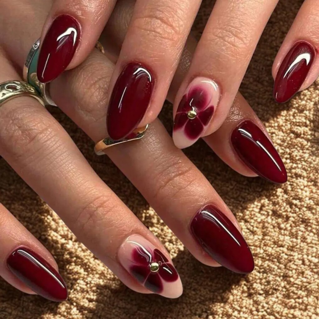 Close-up of manicured hands with pointed burgundy nails, featuring a floral design on one nail and gold accents.