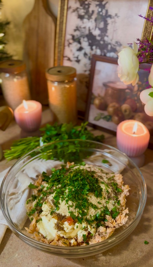 A clear glass bowl filled with a mixture of shredded salmon, bread crumbs, and topped with chopped fresh herbs, placed on a table decorated with candles, jars, and framed pictures.
