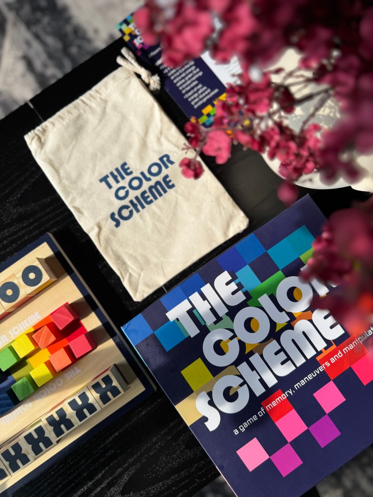 An overhead view of 'The Color Scheme' board game, featuring a canvas storage pouch, colorful wooden blocks, code cubes, and the game box on a dark surface, with flowers slightly blurred in the background.