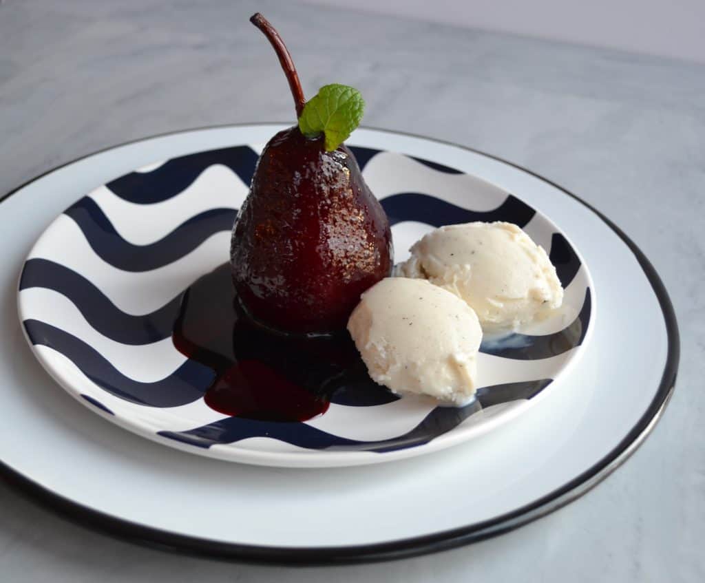 A poached pear glazed in a deep red sauce, served on a wavy patterned plate with two scoops of vanilla ice cream and a sprig of mint for garnish.