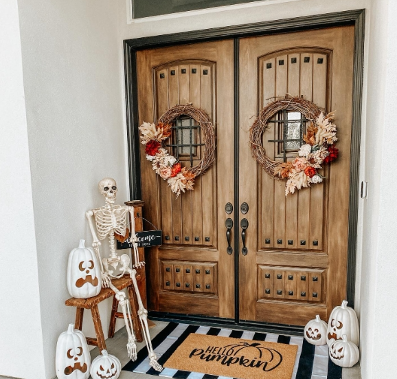 A cozy porch with double wooden doors adorned with autumn wreaths, featuring a skeleton beside a welcome sign and decorative pumpkin accents, creating a playful fall atmosphere.