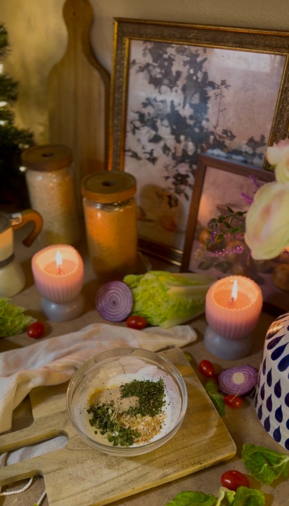 A wooden cutting board with a bowl of spices and herbs, surrounded by candles, decorative jars, fresh vegetables like lettuce and tomatoes, and a vintage framed picture in a cozy kitchen setting.